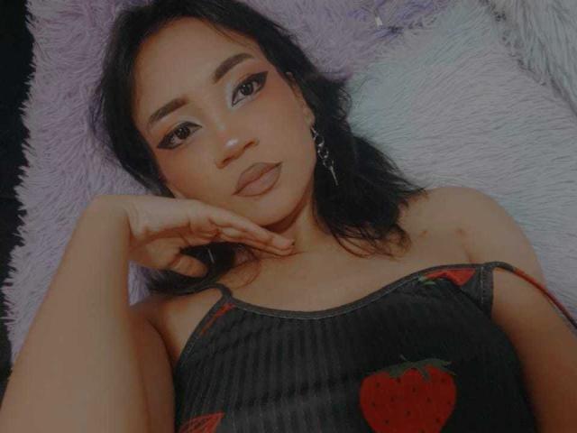 HenutmireRossi deitada em um travesseiro roxo, cabelo preto, olhos escuros, delineador, batom, blusa preta com alças finas e morangos vermelhos, mão embaixo do queixo, olhando para a câmera, luz suave..