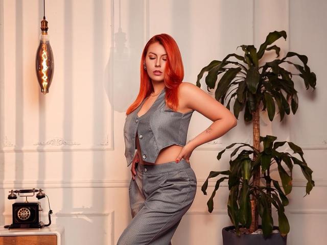 AnnyCraffort with red hair, wearing a gray sleeveless vest and matching pants, standing with hands on hips, showing her tits and ass, next to a potted plant.