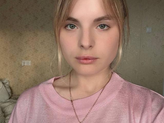 Andrea Filmore hat helgrüne Augen, einen rosafarbenen Pullover und eine zarte Halskette. Sie blickt direkt in die Kamera mit einem neutralen Gesichtsausdruck. Ihr blondes Haar ist zu einem lockeren Pferdeschwanz gebunden..