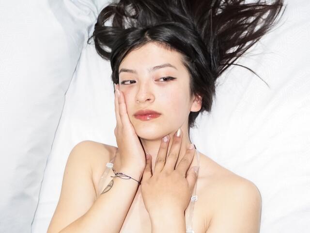 HailyEvans with dark hair, pale skin, and pink eyeliner, lying on white pillows, hands on her face, glossy lips, and a tattoo on her right wrist, topless.