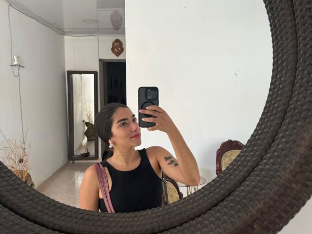 SharonKlart with black tank top, holding phone in mirror selfie, brown hair in bun, small tattoo on bicep, pink strap bag, white wall, dark circular mirror.