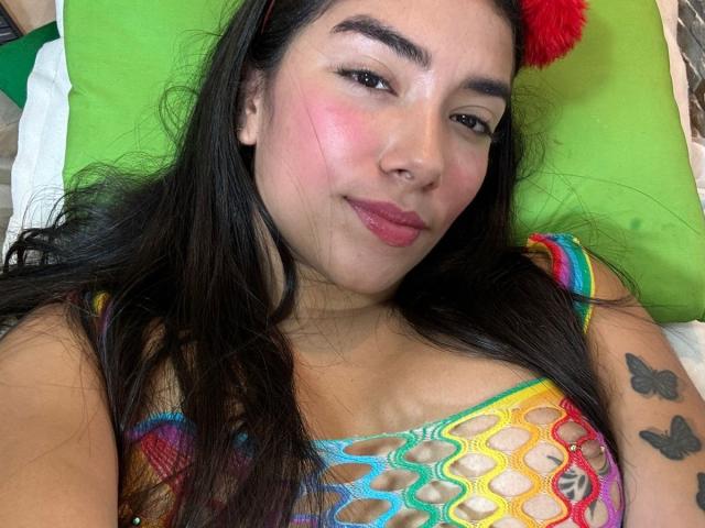 SharonKlart with pink cheeks, long black hair, wearing a rainbow crochet top, showing small tattoos of butterflies on her right arm, lying on a green pillow. Boobs visible.