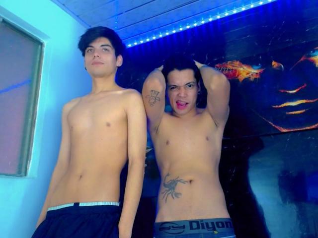 Dois homens sem camisa, TheShiningMen, um com uma tatuagem no braço direito e outra na parte inferior do abdômen, em pé em um quarto com iluminação azul, usando calças pretas..