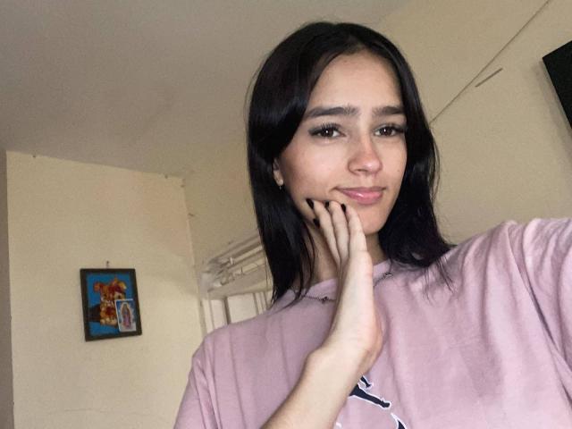 MelodyMansson with black hair, pink shirt, and hand on cheek, smiling subtly, showing cute, small tits, and smooth, fair skin, in a softly lit room.