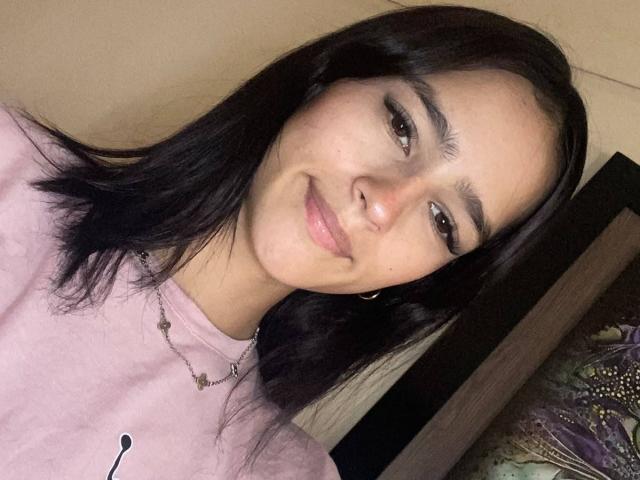 MelodyMansson has dark brown hair, a slight smile, and is wearing a pink shirt with a necklace. She's looking at the camera with a relaxed expression.