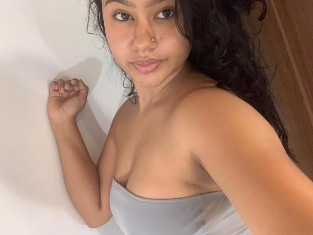 NoiraVale tiene el cabello oscuro y rizado, un piercing en la nariz, y lleva un top gris sin tirantes que deja ver sus senos, y tiene una ligera sonrisa, con el brazo levantado..