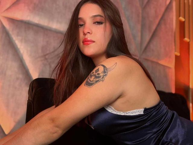 Katarina Novak, mit langen braunen Haaren, rotem Lippenstift und einem blauen Seidenkleid, hat ein Totenkopf-Tattoo auf ihrem rechten Arm und sitzt auf einem schwarzen Samtsessel..