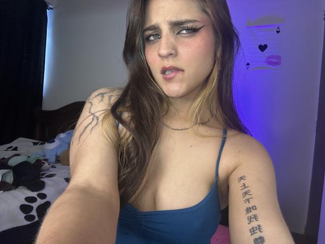 SarahhTanyas with pouty lips, blue tank top, tattoos on arm, brown hair with blonde streaks, eyeliner, nose ring, and a slightly annoyed expression, taking a selfie.