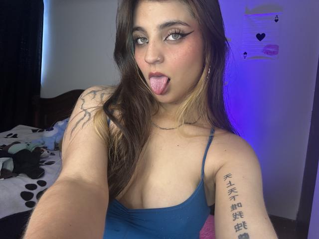 SarahhTanyas with long brown hair, blue tank top, tongue out, tattoo on arm, green eyes, and nose ring, taking a selfie, showing small boobs and a cute face.