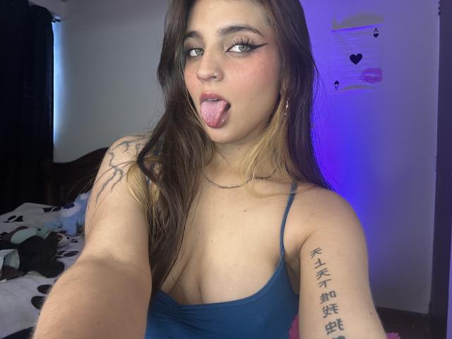 SarahhTanyas with long brown hair, blue tank top, tongue out, tattoos on arm, thick eyeliner, and a cute face, showing off her small boobs and cute ass.
