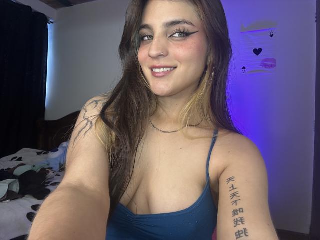 SarahhTanyas with long brown hair, blue tank top, smiling, showing cleavage, tattoo on right arm, makeup with winged eyeliner, in bedroom with blue light. Boobs visible.