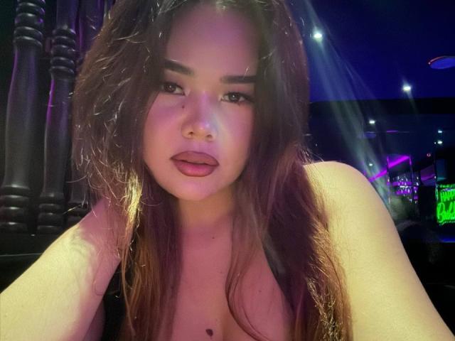 AyaCharm heeft nat bruin haar, volle lippen met roze lippenstift, en een klein sproetje boven haar schaamlippen. Ze is topless, met zichtbare borsten, in een zwak verlichte, blauwige bar..