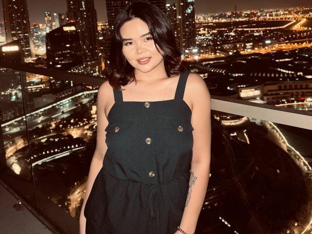 AyaCharm mit mittlerer Brustgröße, in einem schwarzen Kleid mit Knopfleiste, stehend vor einem Glasbalkon in der Nacht, Stadtlichter im Hintergrund, dunkles, welliges Haar, lächelnd, Tattoo am rechten Arm..