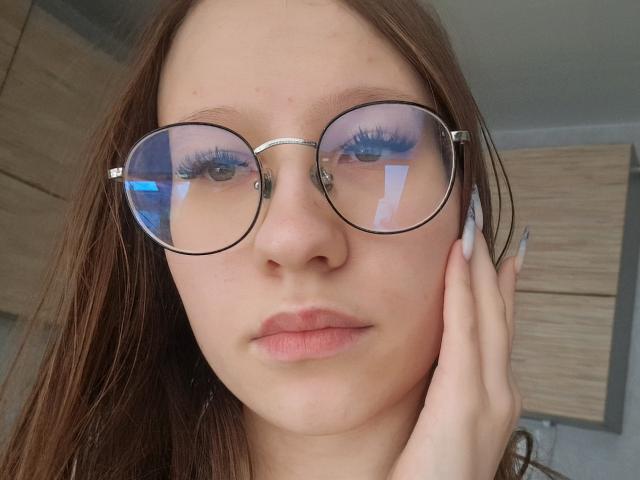 VelvetSinn with round glasses, reflecting light, has brown hair, blue eyes with long lashes, pink lips, and holds her cheek with long, manicured nails. Her pussy isn't visible.