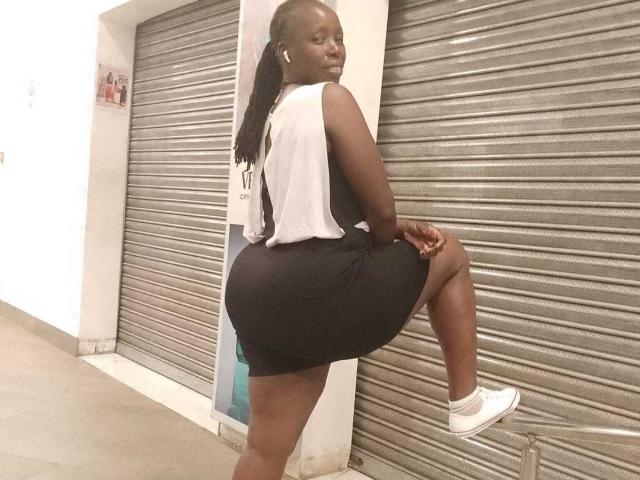 BeutyLicious, dark-skinned, with a curvy ass, wearing a white sleeveless top and black shorts, has one leg raised, showing white sneakers, and wearing earbuds.