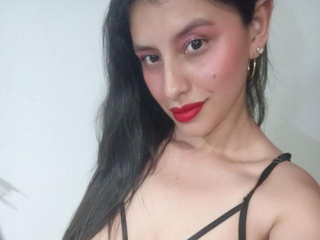 MadissonFontaine with red lipstick, pink eyeshadow, and black strappy top, showing off her tits and earrings, black hair, fair skin, and a cute smile.