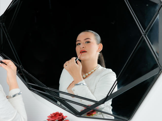 IsabelCarson with slicked-back dark hair, applying red lipstick, wearing a white top, pearl necklace, and white bracelet, reflected in a black, diamond-shaped mirror.