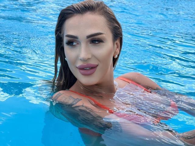 NicolSNaya with wet brown hair, pink lipstick, and tattoos, wearing an orange bikini, smiling in a blue pool, showing her tits above the water.
