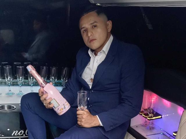 ErosPoomps in een blauw pak, met een roze fles en een glas in de hand, met een serieuze blik en een lichte frons, zittend naast een aantal champagneglazen..
