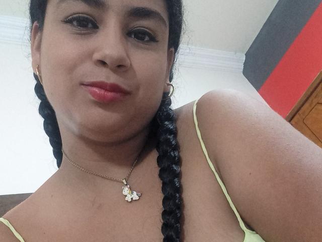 AlisonHank has dark skin, red lipstick, and black braided hair, wearing a green strap top and gold necklace with a butterfly pendant, smiling slightly. Her boobs are visible.