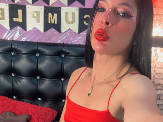 ArielNitzche with red lipstick, black eyeliner, and red tank top, pouting with shiny skin, black hair, and a gold necklace, on a black tufted headboard.