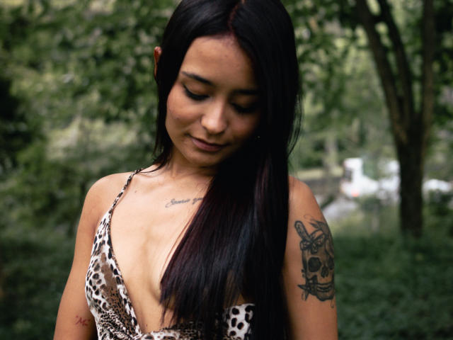 ShantellRuiz with long black hair, looking down, wearing a leopard-print halter top, showing small tits, tattoo on right arm, tattoo on chest, in a green, blurry outdoor setting.