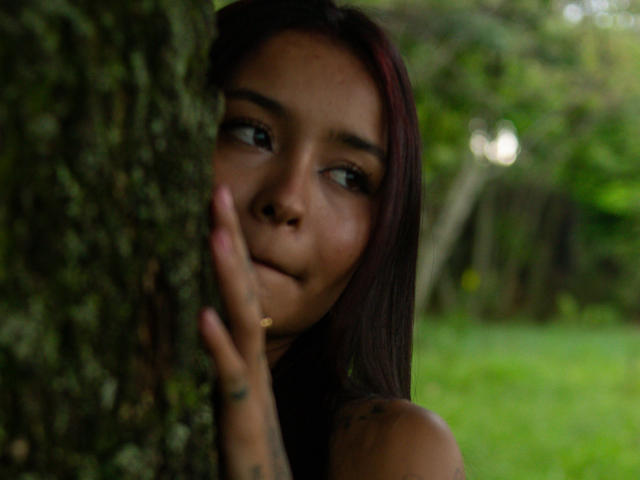 ShantellRuiz met donkerbruin haar, die een boomstam vasthoudt, haar gezicht is half verborgen, ze kijkt opzij, met een lichte glimlach, een tatoeage op haar vinger, een zachte groene achtergrond, natuurlijk licht, subtiele make-up..