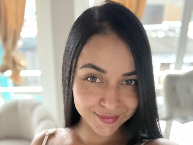 HikariChloe tiene una piel marrón lisa, cabello negro y lacio, ojos marrones y una pequeña sonrisa con lápiz labial rosa. Lleva una blusa blanca, luciendo linda y sexy..