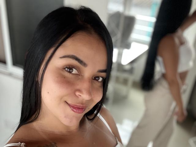 HikariChloe, con cabello negro, ojos marrones y una ligera sonrisa, vestida con una blusa blanca, mostrando un reflejo de sus glúteos en el fondo..