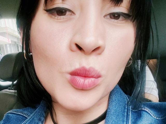 IssisDiosa's face with pouty pink lips, black eyeliner, and straight black hair, wearing a blue denim jacket, inside a car. Her skin is light, and she has small earrings.