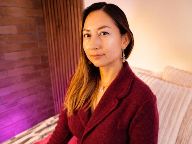 GiaddaHeyan, con piel de color marrón claro, cabello castaño liso, vistiendo un blazer de punto rojo, pequeños aretes y una sonrisa sutil, sentada frente a una pared blanca..