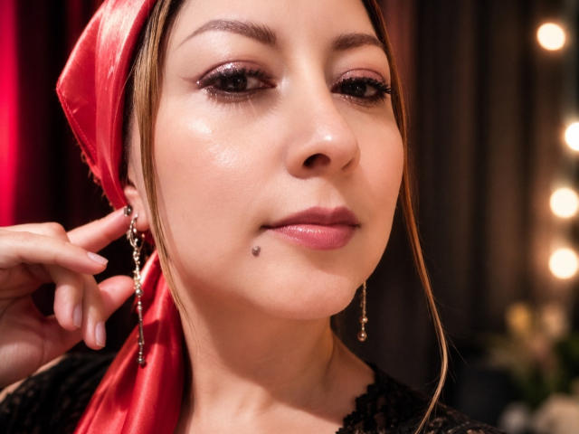 GiaddaHeyan, met een rode hoofddoek, bruine ogen, subtiele make-up, zwarte eyeliner, gouden oorbellen en een klein beauty markje op haar onderlip, met een enigszins melancholische blik..