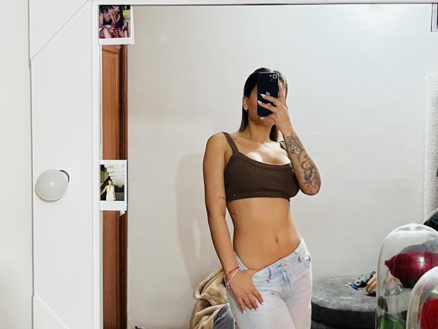 ThaliaAres with brown crop top, showing tits, and tattoos, wearing light blue jeans, taking a mirror selfie, holding a black phone, in a white-walled room.