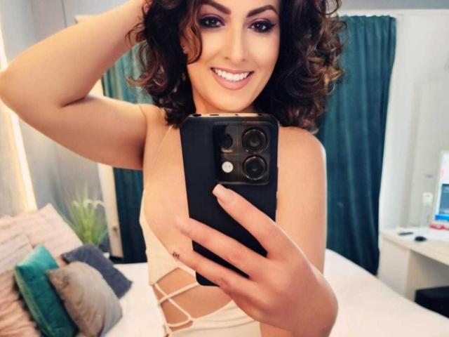 LisaCroftt, met krullend bruin haar, lachend, met een zwarte telefoon in haar hand, die haar borsten laat zien in een witte, open top, die een spiegel-selfie maakt, in een slaapkamer..