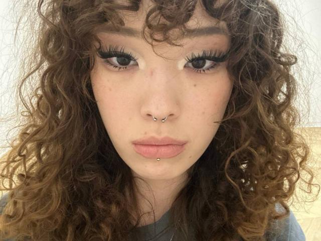 ZolaBouret has curly brown hair, black eyeliner, and a nose ring. She's also sporting a lower lip piercing with a small ball. Her eyes are looking forward.