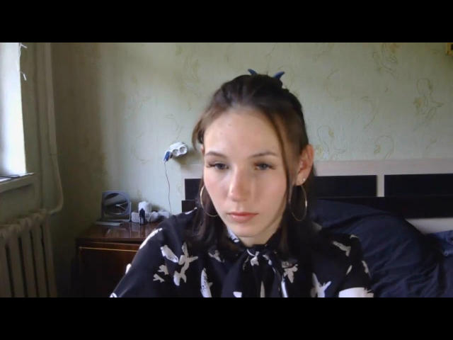 NathalieDutt has pale skin, brown hair in a ponytail, wearing a black shirt with white bird patterns, and large hoop earrings, sitting in a room with a lamp.
