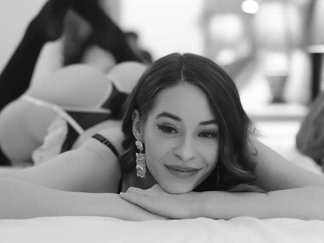 Fotografía en blanco y negro de MissMinna acostada en una cama, boca abajo, sonriendo, con cabello largo y oscuro, usando aretes y un sostén, mostrando sus senos y un vistazo de su trasero..