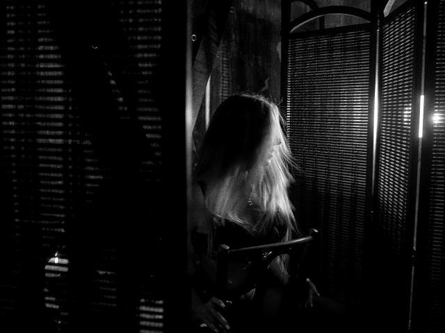 Black-and-white photo of AylinRoze with long hair, looking down, holding a sword, standing in front of a folding screen with slatted patterns, shadowed and mysterious.