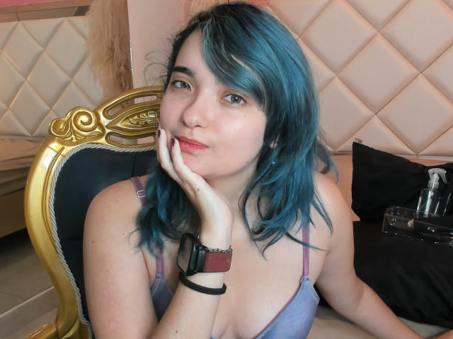 Naommy Carter mit blauen Haaren, heller Haut und einem lila BH, die ihr Kinn in die Hand legt, sieht niedlich mit einem leichten Lächeln aus und trägt ein braunes Armband..