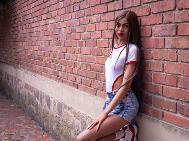Crystal Davids, com cabelos castanhos longos, blusa branca e vermelha e shorts jeans, apoiada em uma parede de tijolos, exibindo suas nádegas em shorts justos, com saltos altos vermelhos..