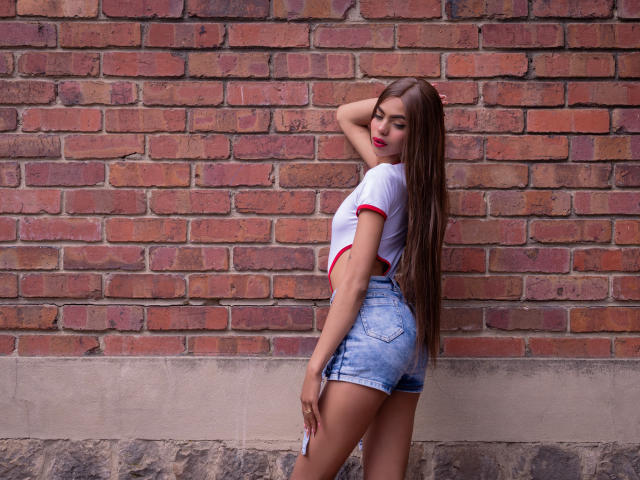 CrystalDavids, avec de longs cheveux bruns, un débardeur blanc à bordure rouge et un short en jean, debout devant un mur de briques, regardant en arrière, une main sur la tête, montrant ses fesses..