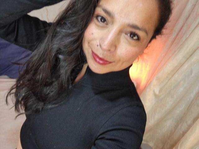StangelarRivera with dark wavy hair, wearing a black turtleneck, smiling with red lipstick, showing a small nose boop, and holding a purple garment on the left.
