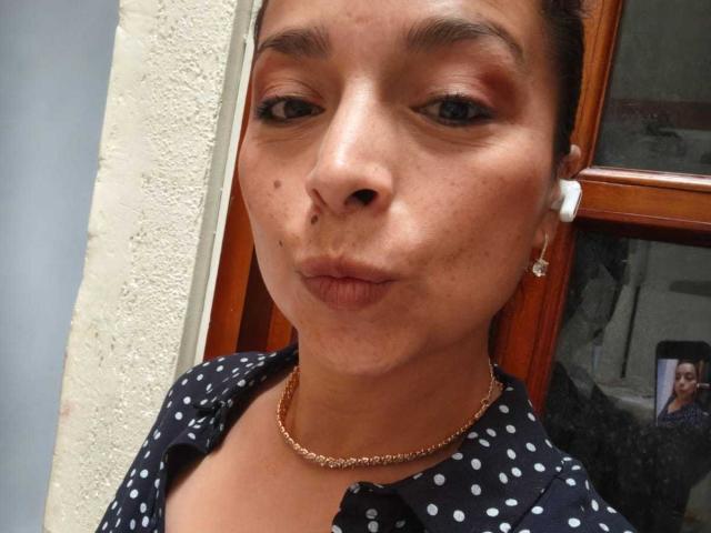 StangelarRivera with puffy lips, brown skin, small nose, polka dot shirt, gold chain, earrings, and white earbud in a selfie. She's making a pouty face with slightly raised eyebrows.