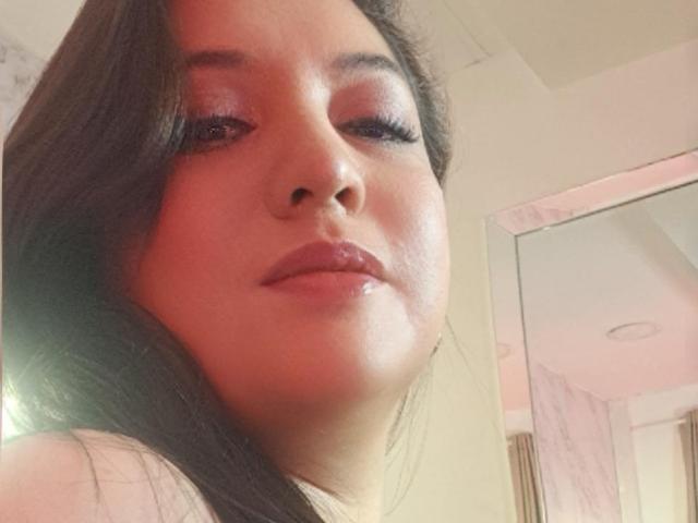PaulaAnd has shiny pink eyeshadow, long dark eyelashes, glossy pink lips, and brown wavy hair. Her face is close to the camera with a slight, confident smile.