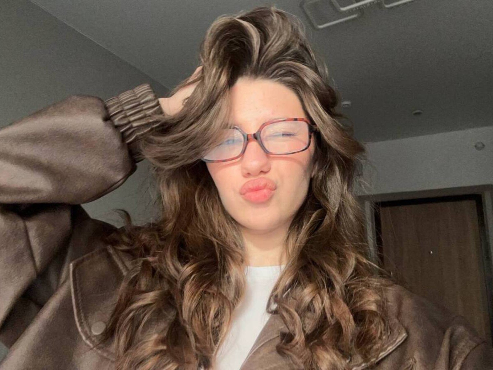 Karisa Stathas, con el pelo castaño ondulado, usando gafas y una chaqueta de cuero marrón, frunciendo sus labios rosados. Está tocándose el pelo con una mano, y se ve linda..