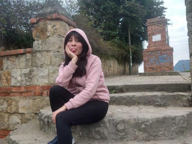 IssisDiosa, with puffy pink hoodie and black pants, sits on stone steps, hand on cheek, looking cute, her ass slightly visible, in a rustic, brick-walled setting.