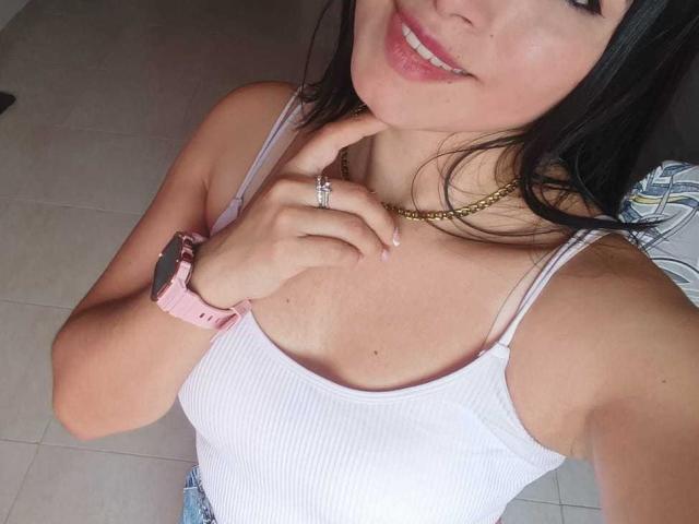 AlexaLaker mit mittelgroßer Brust, in einem weißen, gerippten Tanktop, lächelnd, mit einer rosa Armbanduhr, einer goldenen Halskette, einem Fingerring und schwarzen Haaren, stehend auf einem gefliesten Boden..