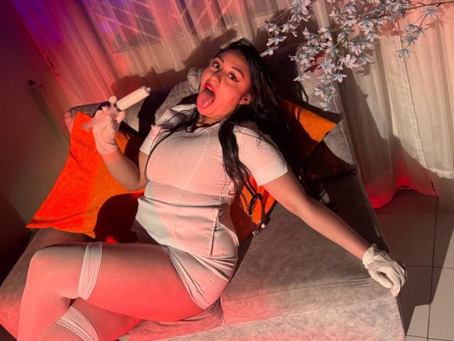 CristalZafiro, com longos cabelos pretos, língua de fora, vestindo um vestido branco justo, luvas brancas e faixas nas coxas, segurando um bastão branco, sentada em um sofá cinza com almofadas laranja..
