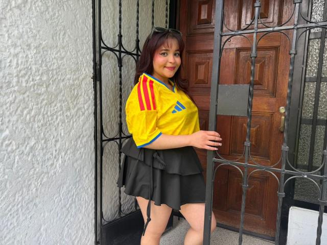 SamyMancini, with red-brown hair, wearing a yellow soccer jersey with red stripes and black skirt, standing by a metal gate, smiling at the camera.