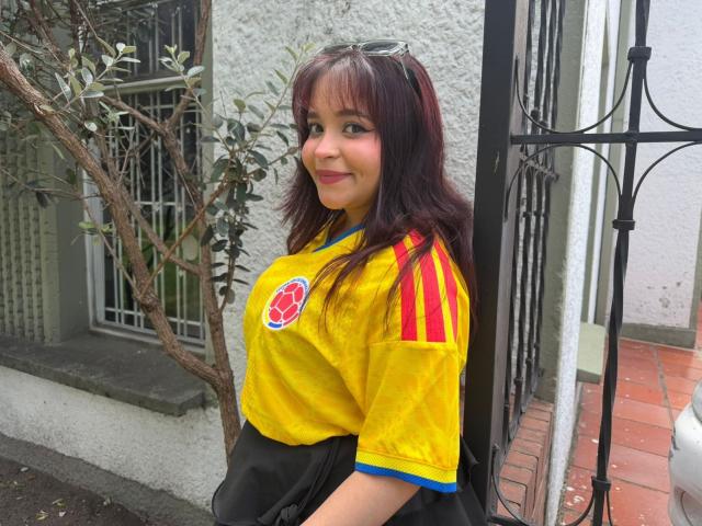 SamyMancini with medium tits in a yellow soccer shirt with red stripes, black skirt, and brown hair, standing by a black metal gate, smiling.